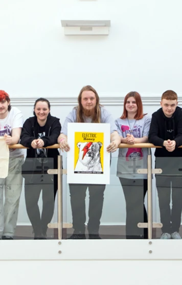 Group of music business students and a lecturer lined up along a balcony, some holding Electric Honey tote bags and a framed anniversary poster. Group of music business students and a lecturer lined up along a balcony, some holding Electric Honey tote bags and a framed anniversary poster.